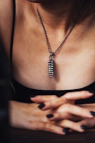 A person wearing a metallic pendant on a beaded chain around their neck. The pendant is elongated and textured, resembling a series of stacked segments. The individual's hands are crossed in front, with fingers interlaced. The lighting creates a moody, intimate atmosphere, highlighting the textures and contours of the skin and jewelry.