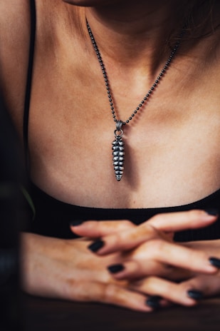 A person wearing a metallic pendant on a beaded chain around their neck. The pendant is elongated and textured, resembling a series of stacked segments. The individual's hands are crossed in front, with fingers interlaced. The lighting creates a moody, intimate atmosphere, highlighting the textures and contours of the skin and jewelry.