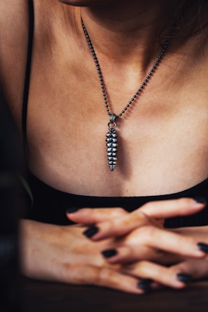 A person wearing a metallic pendant on a beaded chain around their neck. The pendant is elongated and textured, resembling a series of stacked segments. The individual's hands are crossed in front, with fingers interlaced. The lighting creates a moody, intimate atmosphere, highlighting the textures and contours of the skin and jewelry.