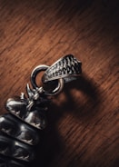 A close-up photograph of a metallic object with intricate detailing, placed on a wooden surface. The object appears to be a piece of jewelry, possibly a pendant or part of an accessory, with a textured finish.