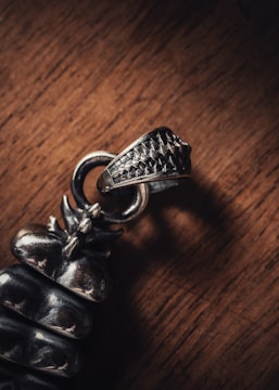 A close-up photograph of a metallic object with intricate detailing, placed on a wooden surface. The object appears to be a piece of jewelry, possibly a pendant or part of an accessory, with a textured finish.