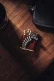 A detailed view of a metal and leather lighter on a wooden surface, featuring a unique design with metallic finger-like elements gripping the body. The lighting creates a warm, rustic ambiance.