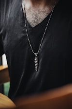 A person wearing a black V-neck shirt with a long necklace that features a spiral pendant. The chest hair is visible, and there is a blurred background.