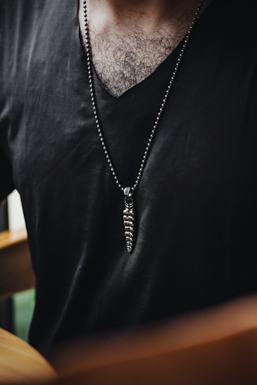 A person wearing a black V-neck shirt with a long necklace that features a spiral pendant. The chest hair is visible, and there is a blurred background.