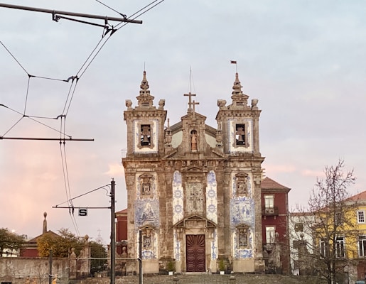 A historic church with two bell towers stands in the foreground, adorned with blue and white azulejos tiles. The sky is softly lit by the setting or rising sun, casting a warm glow. Overhead cables from an electric tram system are visible, emphasizing the urban setting.