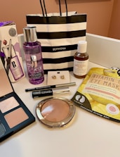 An arrangement of various cosmetic and skincare products displayed, including Clinique makeup remover, an eyeshadow palette, Kiehl's cleanser, vitamin eye mask, a pair of earrings, and several other makeup items. A black and white striped Sephora shopping bag is also present.