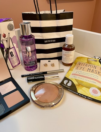 An arrangement of various cosmetic and skincare products displayed, including Clinique makeup remover, an eyeshadow palette, Kiehl's cleanser, vitamin eye mask, a pair of earrings, and several other makeup items. A black and white striped Sephora shopping bag is also present.
