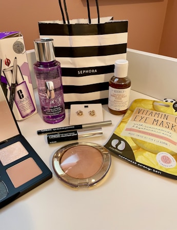 An arrangement of various cosmetic and skincare products displayed, including Clinique makeup remover, an eyeshadow palette, Kiehl's cleanser, vitamin eye mask, a pair of earrings, and several other makeup items. A black and white striped Sephora shopping bag is also present.