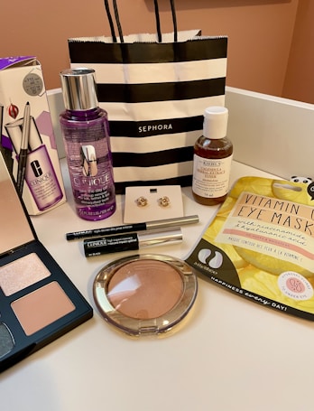 An arrangement of various cosmetic and skincare products displayed, including Clinique makeup remover, an eyeshadow palette, Kiehl's cleanser, vitamin eye mask, a pair of earrings, and several other makeup items. A black and white striped Sephora shopping bag is also present.
