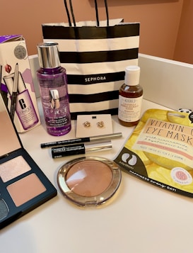 An arrangement of various cosmetic and skincare products displayed, including Clinique makeup remover, an eyeshadow palette, Kiehl's cleanser, vitamin eye mask, a pair of earrings, and several other makeup items. A black and white striped Sephora shopping bag is also present.