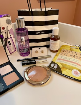 An arrangement of various cosmetic and skincare products displayed, including Clinique makeup remover, an eyeshadow palette, Kiehl's cleanser, vitamin eye mask, a pair of earrings, and several other makeup items. A black and white striped Sephora shopping bag is also present.