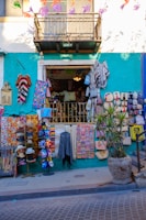 A colorful street shop with various handmade items and textiles displayed outside. The facade is painted in a bright teal with a white upper level. Items include vibrant blankets, bags, masks, and hats. Decorative banners hang above, and a potted plant is placed to the right.