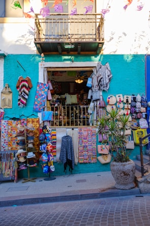 A colorful street shop with various handmade items and textiles displayed outside. The facade is painted in a bright teal with a white upper level. Items include vibrant blankets, bags, masks, and hats. Decorative banners hang above, and a potted plant is placed to the right.