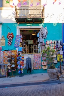 A colorful street shop with various handmade items and textiles displayed outside. The facade is painted in a bright teal with a white upper level. Items include vibrant blankets, bags, masks, and hats. Decorative banners hang above, and a potted plant is placed to the right.