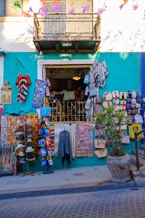A colorful street shop with various handmade items and textiles displayed outside. The facade is painted in a bright teal with a white upper level. Items include vibrant blankets, bags, masks, and hats. Decorative banners hang above, and a potted plant is placed to the right.