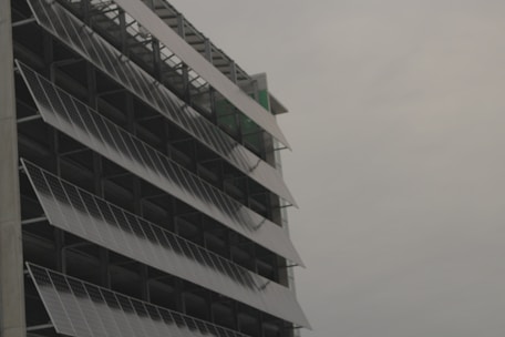 A modern building facade with rows of tilted solar panels mounted along the structure, set against a cloudy sky.