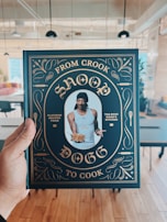 A person is holding a cookbook titled 'From Crook to Cook' by Snoop Dogg in a stylish cover featuring gold ornate designs on a dark background. The central image on the cover shows a man wearing a tank top and a black head wrap, holding a plate of food. The setting appears to be a modern, open-plan room with tables and chairs.