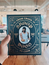 A person is holding a cookbook titled 'From Crook to Cook' by Snoop Dogg in a stylish cover featuring gold ornate designs on a dark background. The central image on the cover shows a man wearing a tank top and a black head wrap, holding a plate of food. The setting appears to be a modern, open-plan room with tables and chairs.