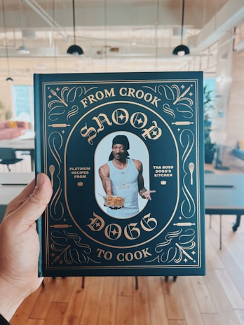A person is holding a cookbook titled 'From Crook to Cook' by Snoop Dogg in a stylish cover featuring gold ornate designs on a dark background. The central image on the cover shows a man wearing a tank top and a black head wrap, holding a plate of food. The setting appears to be a modern, open-plan room with tables and chairs.