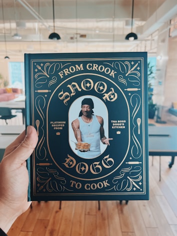 A person is holding a cookbook titled 'From Crook to Cook' by Snoop Dogg in a stylish cover featuring gold ornate designs on a dark background. The central image on the cover shows a man wearing a tank top and a black head wrap, holding a plate of food. The setting appears to be a modern, open-plan room with tables and chairs.