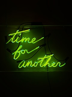 Neon green lettering spells out the phrase 'time for another' against a dark background. The glowing letters are stylishly curved and illuminated, creating a striking and vibrant visual effect.