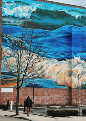 A massive mural depicting vibrant ocean waves covers the side of a brick building. In the foreground, a person dressed in dark clothing walks next to leafless trees on the sidewalk. The artwork on the building is rich in blue hues and white foam, capturing the dynamic movement of waves.