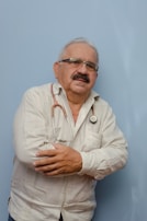An older man with glasses and a mustache is wearing a light-colored, long-sleeved shirt. A stethoscope is around his neck, and he has his arms crossed. The background is a solid light blue color.