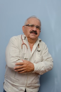 An older man with glasses and a mustache is wearing a light-colored, long-sleeved shirt. A stethoscope is around his neck, and he has his arms crossed. The background is a solid light blue color.