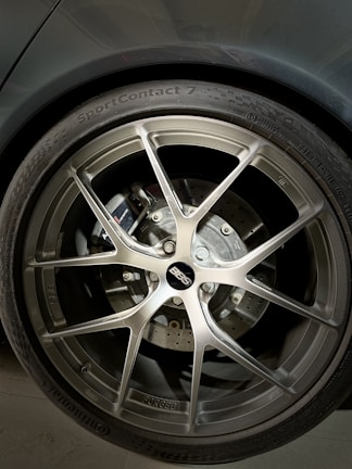 A close-up of a car wheel featuring a BBS alloy rim, with a visible brake disc and caliper behind it. The tire is labeled with 'SportContact 7' by Continental, indicating a high-performance tire. The metallic finish of the rim contrasts with the darker tones of the tire and the car body.