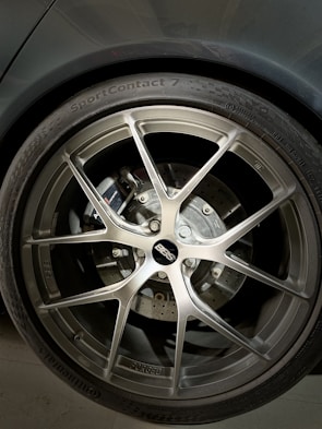 A close-up of a car wheel featuring a BBS alloy rim, with a visible brake disc and caliper behind it. The tire is labeled with 'SportContact 7' by Continental, indicating a high-performance tire. The metallic finish of the rim contrasts with the darker tones of the tire and the car body.