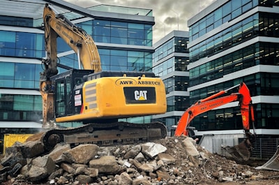 An excavator is placed on top of a pile of rubble amidst a construction site. The construction equipment is positioned in an urban environment, surrounded by modern glass office buildings. The machinery is labeled with the brand 'Caterpillar' and the name 'AWR Abbruch'. A second orange excavator stands nearby, contributing to the ongoing demolition work. The atmosphere suggests a busy site as the excavators appear functional and operational.