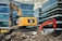 An excavator is placed on top of a pile of rubble amidst a construction site. The construction equipment is positioned in an urban environment, surrounded by modern glass office buildings. The machinery is labeled with the brand 'Caterpillar' and the name 'AWR Abbruch'. A second orange excavator stands nearby, contributing to the ongoing demolition work. The atmosphere suggests a busy site as the excavators appear functional and operational.