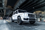A white luxury SUV is parked under a dimly lit urban overpass. The car features a sleek, modern design with large wheels and a distinctive grille. Overhead, concrete structures and beams are illuminated by spotlights, casting shadows on the roadway. Traffic cones and yellow tape are visible in the background near a wall.