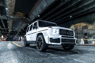 A white luxury SUV is parked under a dimly lit urban overpass. The car features a sleek, modern design with large wheels and a distinctive grille. Overhead, concrete structures and beams are illuminated by spotlights, casting shadows on the roadway. Traffic cones and yellow tape are visible in the background near a wall.