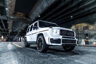 A white luxury SUV is parked under a dimly lit urban overpass. The car features a sleek, modern design with large wheels and a distinctive grille. Overhead, concrete structures and beams are illuminated by spotlights, casting shadows on the roadway. Traffic cones and yellow tape are visible in the background near a wall.