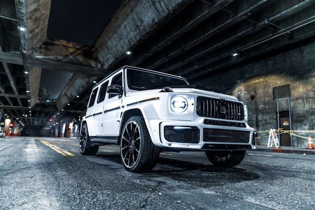 A white luxury SUV is parked under a dimly lit urban overpass. The car features a sleek, modern design with large wheels and a distinctive grille. Overhead, concrete structures and beams are illuminated by spotlights, casting shadows on the roadway. Traffic cones and yellow tape are visible in the background near a wall.