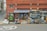A small local store situated in a brick building with a striped blue and white awning. The storefront displays various items in the window. An old-fashioned telephone booth stands to the left, painted green. Several chairs and miscellaneous objects like traffic cones and cleaning supplies are placed outside.