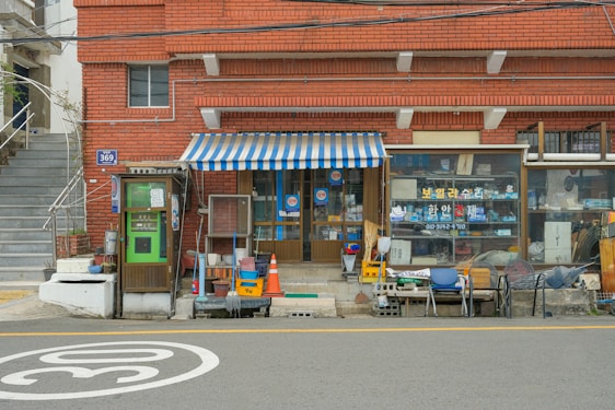 A small local store situated in a brick building with a striped blue and white awning. The storefront displays various items in the window. An old-fashioned telephone booth stands to the left, painted green. Several chairs and miscellaneous objects like traffic cones and cleaning supplies are placed outside.