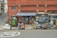 A small local store situated in a brick building with a striped blue and white awning. The storefront displays various items in the window. An old-fashioned telephone booth stands to the left, painted green. Several chairs and miscellaneous objects like traffic cones and cleaning supplies are placed outside.
