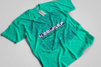 A turquoise t-shirt with an intricate blue graphic design and text on the front. The design includes various abstract shapes, musical elements, and patterns. The text reads 'New Breed of Deep House' with additional words above it that are difficult to discern.