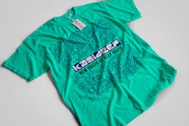A turquoise t-shirt with an intricate blue graphic design and text on the front. The design includes various abstract shapes, musical elements, and patterns. The text reads 'New Breed of Deep House' with additional words above it that are difficult to discern.