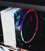 A close-up view of a computer cooling system featuring a large, illuminated fan with vibrant RGB lighting. The blades of the fan are surrounded by a circular strip of colorful LED lights, displaying a spectrum of colors including red, blue, and green. The cooling system is mounted in a black computer case, and the silver heat sink is visible alongside the fan.