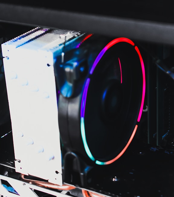 A close-up view of a computer cooling system featuring a large, illuminated fan with vibrant RGB lighting. The blades of the fan are surrounded by a circular strip of colorful LED lights, displaying a spectrum of colors including red, blue, and green. The cooling system is mounted in a black computer case, and the silver heat sink is visible alongside the fan.