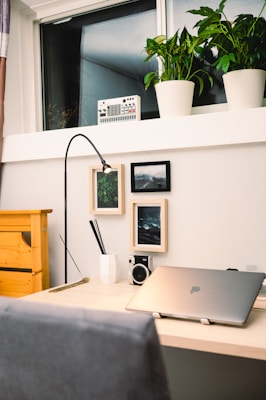 A modern workspace featuring a wooden desk with a laptop, a sleek black lamp, and a small camera. Two framed pictures are hung on the wall above the desk. A white vase containing black pens and a ruler adds to the minimalistic decor. There are several green plants in white pots placed on a shelf by the window, providing a fresh, natural element. Additionally, there is an electronic device on the shelf, possibly an audio mixer.