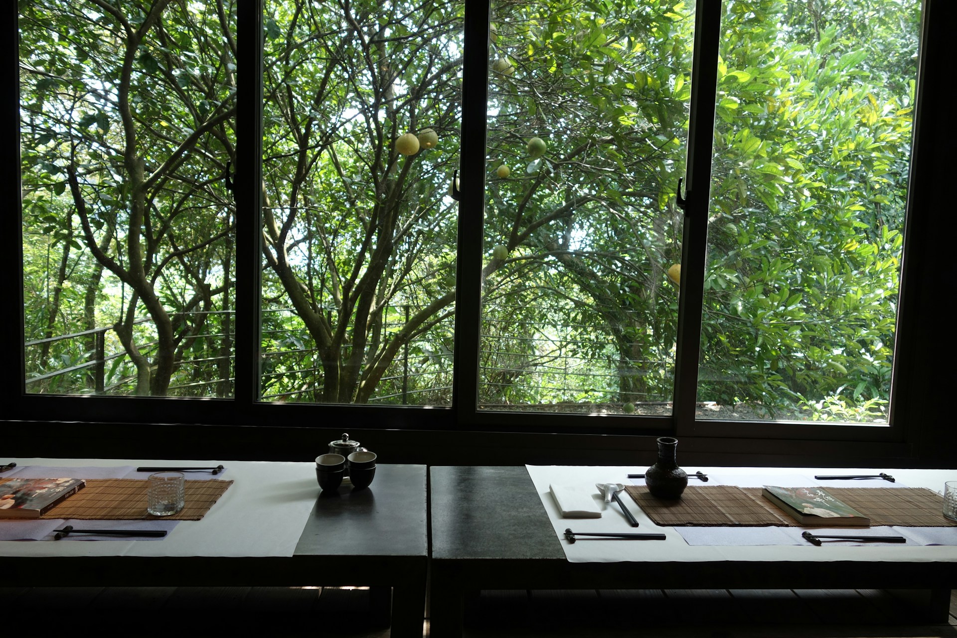 A table is set for dining, with placemats, chopsticks, and small dining vessels. The setting is minimalistic and elegant, with a view of lush green foliage outside a large window. Sunlight filters through the leaves, creating a serene and natural atmosphere.