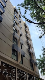 A multi-story building with modern architecture features numerous windows with unique external shading structures. The building's facade is light tan, accentuated by dark window frames. Green trees with lush leaves appear in the foreground and background, against a blue sky.