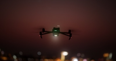 A drone is silhouetted against a dusky sky, its lights glowing faintly in the dimming light. The background is blurred, suggesting a cityscape with scattered lights providing a bokeh effect.