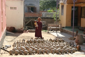 An elderly person dressed in traditional attire stands outdoors holding a child. In front of them is a large collection of earthen pots and pottery arranged neatly on the ground. The setting appears to be a courtyard with buildings on either side. A shadow is cast on the ground, indicating the direction of the sunlight.