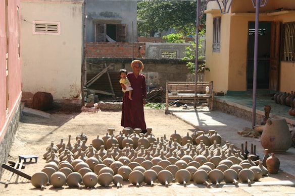 An elderly person dressed in traditional attire stands outdoors holding a child. In front of them is a large collection of earthen pots and pottery arranged neatly on the ground. The setting appears to be a courtyard with buildings on either side. A shadow is cast on the ground, indicating the direction of the sunlight.