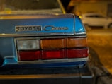 Close-up view of the rear section of a blue vintage Toyota Corona car, showing its badge and taillights. In the background, another car is slightly visible, parked on a dimly lit street.